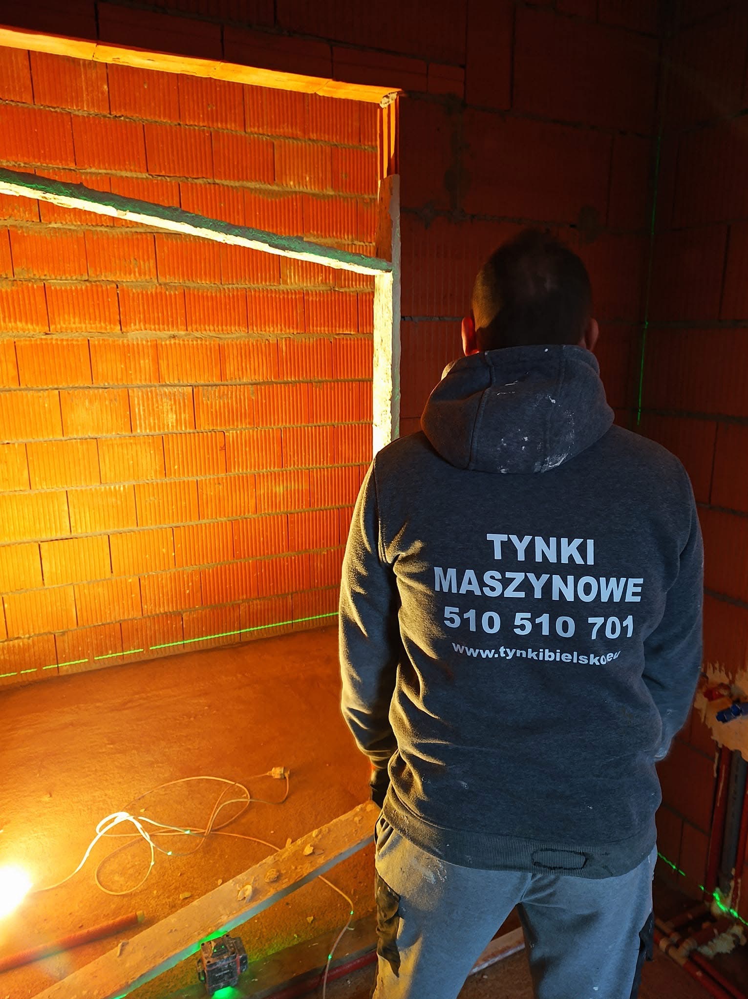 Wnętrze w trakcie prac murarskich z widocznym laserowym niwelatorem oraz pracownikiem w bluzie z nadrukiem firmowym