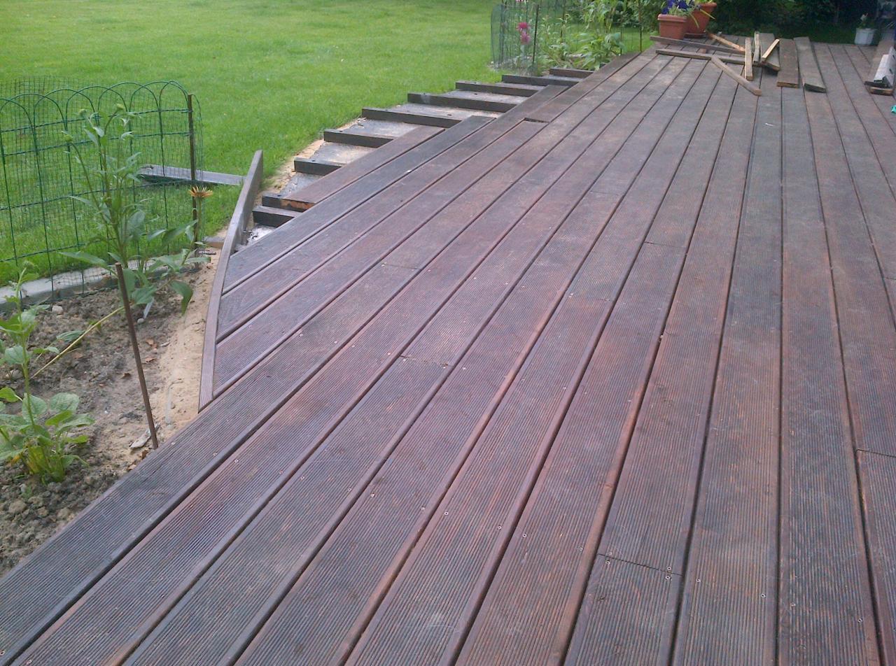 Drewniany taras w ogrodzie, częściowo ukończony, z widocznymi deskami i elementami konstrukcyjnymi. Fragment trawnika i ogrodzenia w tle.