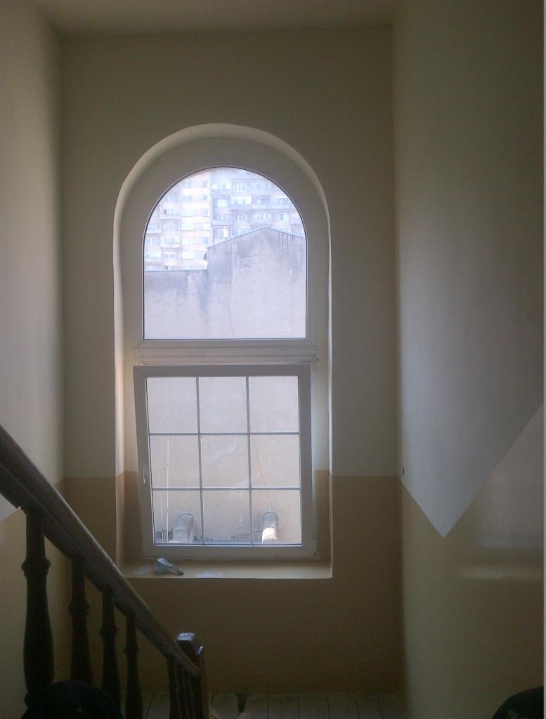 Klatka schodowa po malowaniu z oknem łukowym. Ściany dwukolorowe, odcięcie ukośne. Widok na budynki przez okno z białymi szprosami.