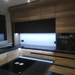 Nowoczesna kuchnia z drewnianymi frontami szafek, czarnymi elementami i wyspą z czarnym, błyszczącym blatem, oświetlenie LED pod szafkami.