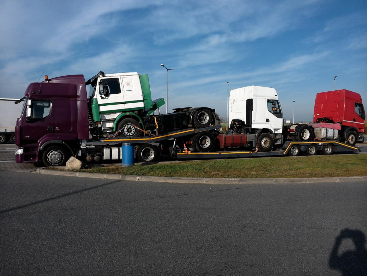 Laweta transportuje trzy kabiny ciężarówek w różnych kolorach: bordowym, białym i czerwonym. Niebieska beczka zabezpiecza ładunek. Widoczny cień osoby.