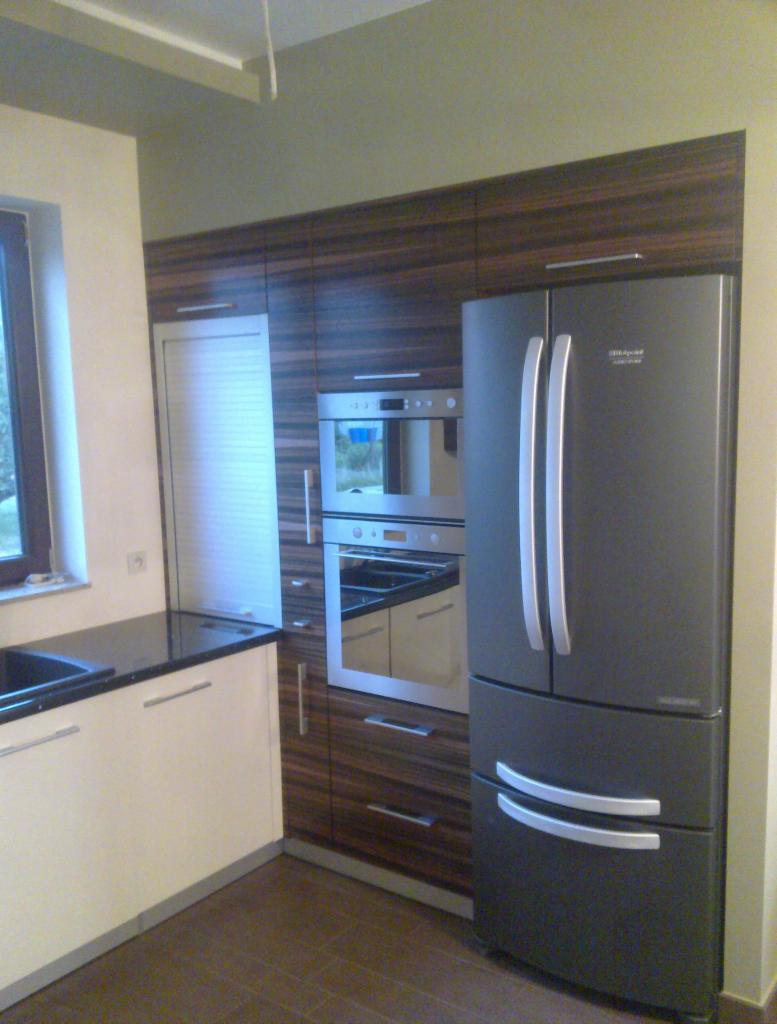 Nowoczesna kuchnia na wymiar z ciemnobrązowymi frontami szafek z widocznym usłojeniem drewna, białymi dolnymi szafkami, czarnym blatem, piekarnikiem w zabudowie i dużą, szarą lodówką side-by-side.