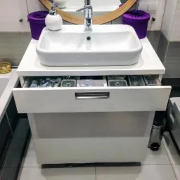 Biała, ceramiczna umywalka nablatowa na szafce z wysuwaną szufladą, w nowoczesnej łazience z szaro-białymi płytkami. Widoczny fragment lustra i akcesoria.