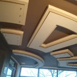 Dekoracyjny sufit podwieszany z geometrycznymi wzorami, oświetleniem punktowym i subtelną kolorystyką, widoczny z perspektywy wnętrza z oknami.