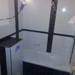 Wykończona łazienka z biało-czarnymi płytkami, widoczna wanna, zabudowa stelaża WC z blatem z mozaiki, lampa halogenowa na kablu.