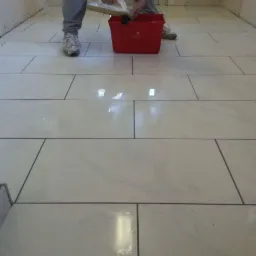 Układanie jasnych, prostokątnych płytek podłogowych z ciemną fugą; widoczny fragment ściany, osoba z narzędziem do fugowania i czerwone wiadro.