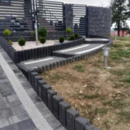 Ułożona szara kostka brukowa z białym pasem, tworząca ścieżkę w ogrodzie, obrzeża z betonowych palisad, w tle nowoczesne ogrodzenie z poziomych paneli i gabionów.