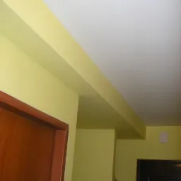 Ściana w odcieniach żółci i bieli po malowaniu, widoczny fragment ościeżnicy z drewnianymi drzwiami i czarne drzwi w tle. Oświetlenie górne.