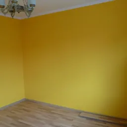 Pomalowane na żółto ściany w pokoju z drewnianą podłogą, widoczny fragment zielonej ściany po prawej stronie i ozdobny żyrandol pod sufitem.
