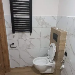 Nowoczesna łazienka z wiszącą toaletą zabudowaną w drewnopodobnej obudowie, białe marmurowe płytki na ścianach, drewnopodobna podłoga, czarny grzejnik drabinkowy i uchwyt na papier toaletowy.