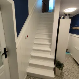 Białe schody po remoncie, z widoczną puszką farby Dulux na szafce, ciemnoniebieska ściana w tle, wzorzysta podłoga i roślina doniczkowa