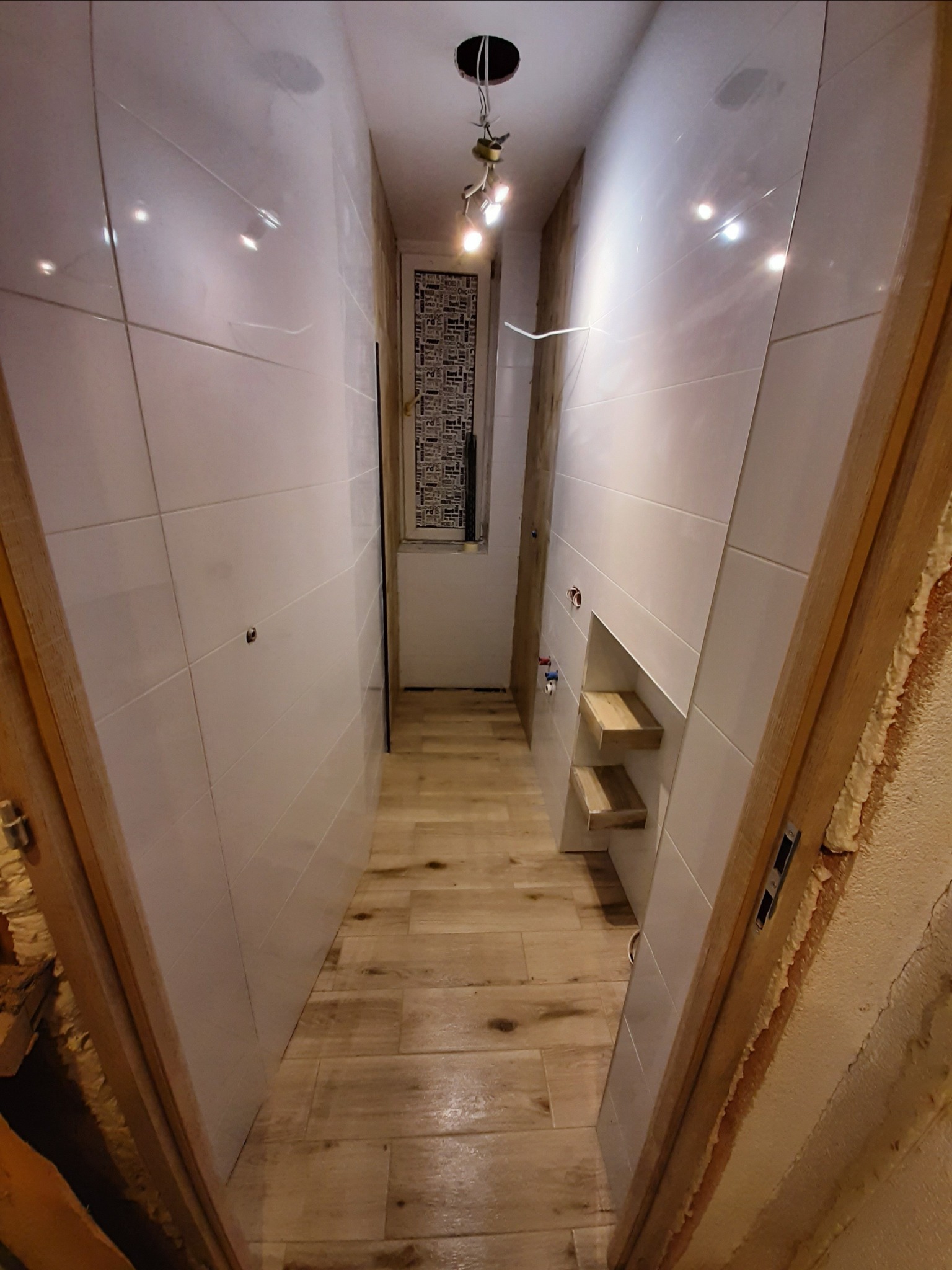 Wąskie wnętrze łazienki w trakcie remontu, z białymi, błyszczącymi płytkami na ścianach, drewnopodobną podłogą, przygotowanymi wnękami na półki i instalacją elektryczną zwisającą z sufitu, widziane...