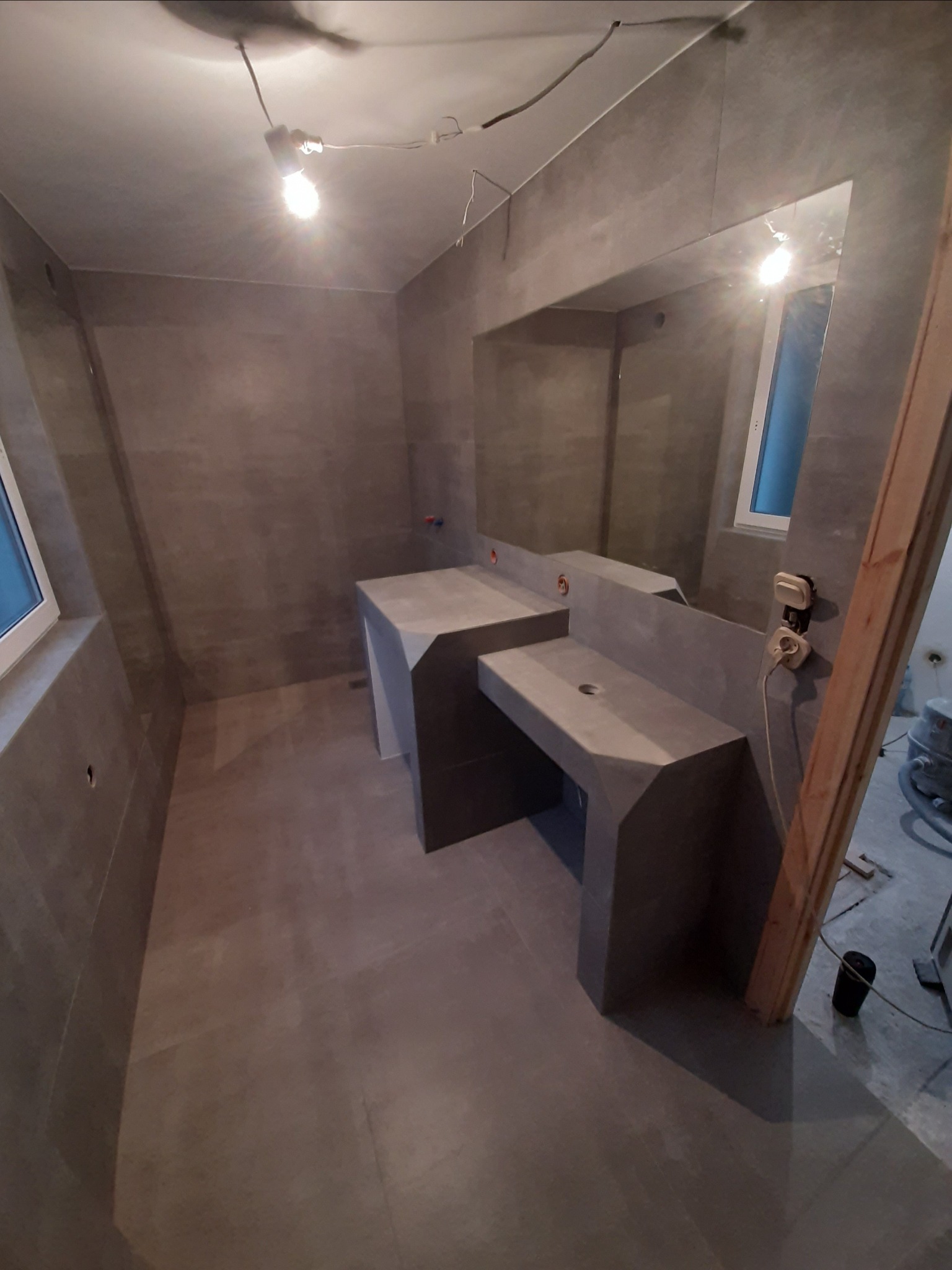 Surowa łazienka w odcieniach szarości z betonowymi umywalkami, widoczne niedokończone prace i prowizoryczne oświetlenie.