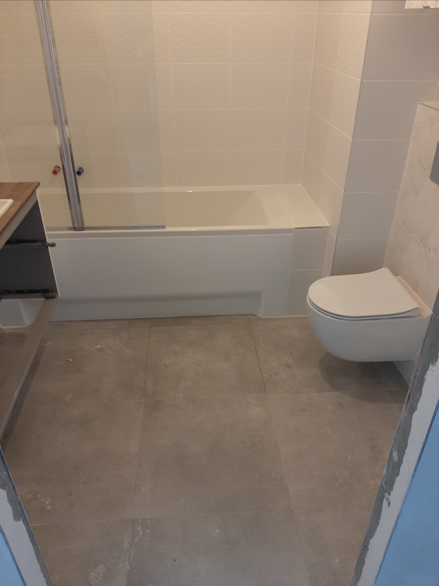 Wyremontowana łazienka z beżowymi płytkami podłogowymi, białą wanną z parawanem nawannowym i podwieszanym WC.