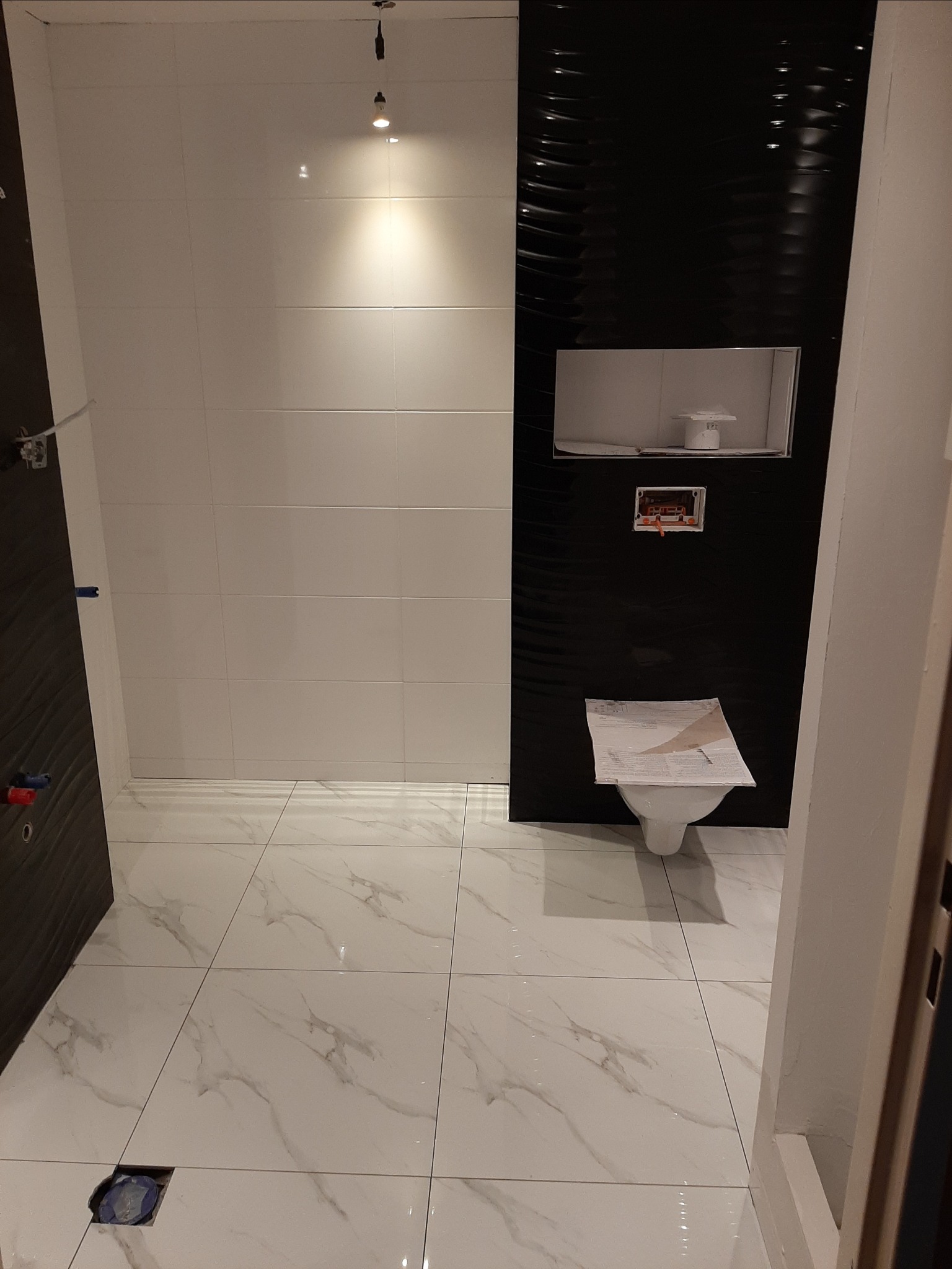 Wnętrze łazienki w trakcie wykańczania: białe płytki na ścianach kontrastują z czarną, falistą ścianą z zabudowanym stelażem WC, na podłodze białe płytki imitujące marmur, przykryta papierem...