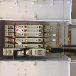 Otwarta skrzynka elektryczna z zamontowanymi bezpiecznikami i przewodami. Widoczne miedziane szyny i etykiety na elementach. Skrzynka w kolorze białym.