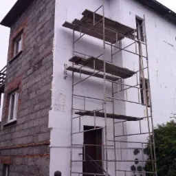 Dom w trakcie ocieplania: połowa elewacji z cegły, druga pokryta styropianem, rusztowanie na tle elewacji, widoczna osoba obserwująca prace.