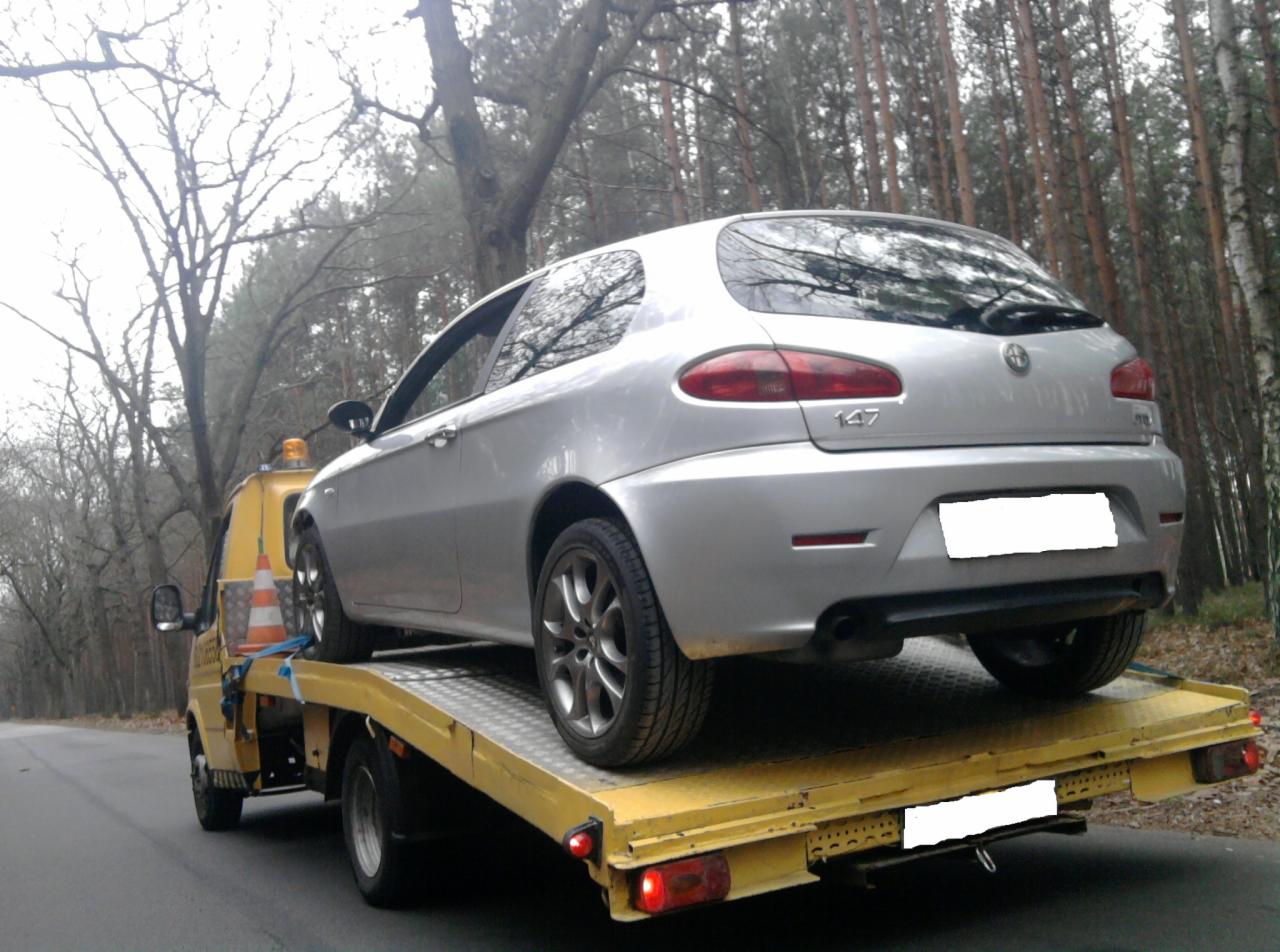 Srebrna Alfa Romeo 147 na żółtej lawecie, widok z tyłu, las w tle. Pomarańczowy pachołek na platformie. Awaryjne światło na dachu lawety.