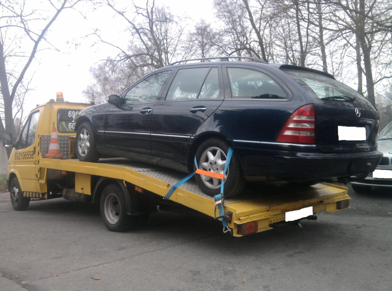 Żółta laweta z niebieskim Mercedesem kombi, zabezpieczonym pasami transportowymi, na platformie. Widoczne drzewa w tle.
