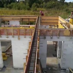 Wznoszenie ścian z bloczków, widoczna szalunkowa konstrukcja ze zbrojeniem, dwóch pracowników na budowie. Budowa domu w trakcie realizacji, widok z góry.