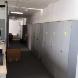 Wnętrze biura po przeprowadzce, widoczne szafy biurowe wzdłuż korytarza, kartony i sprzęt biurowy na biurku po lewej stronie.