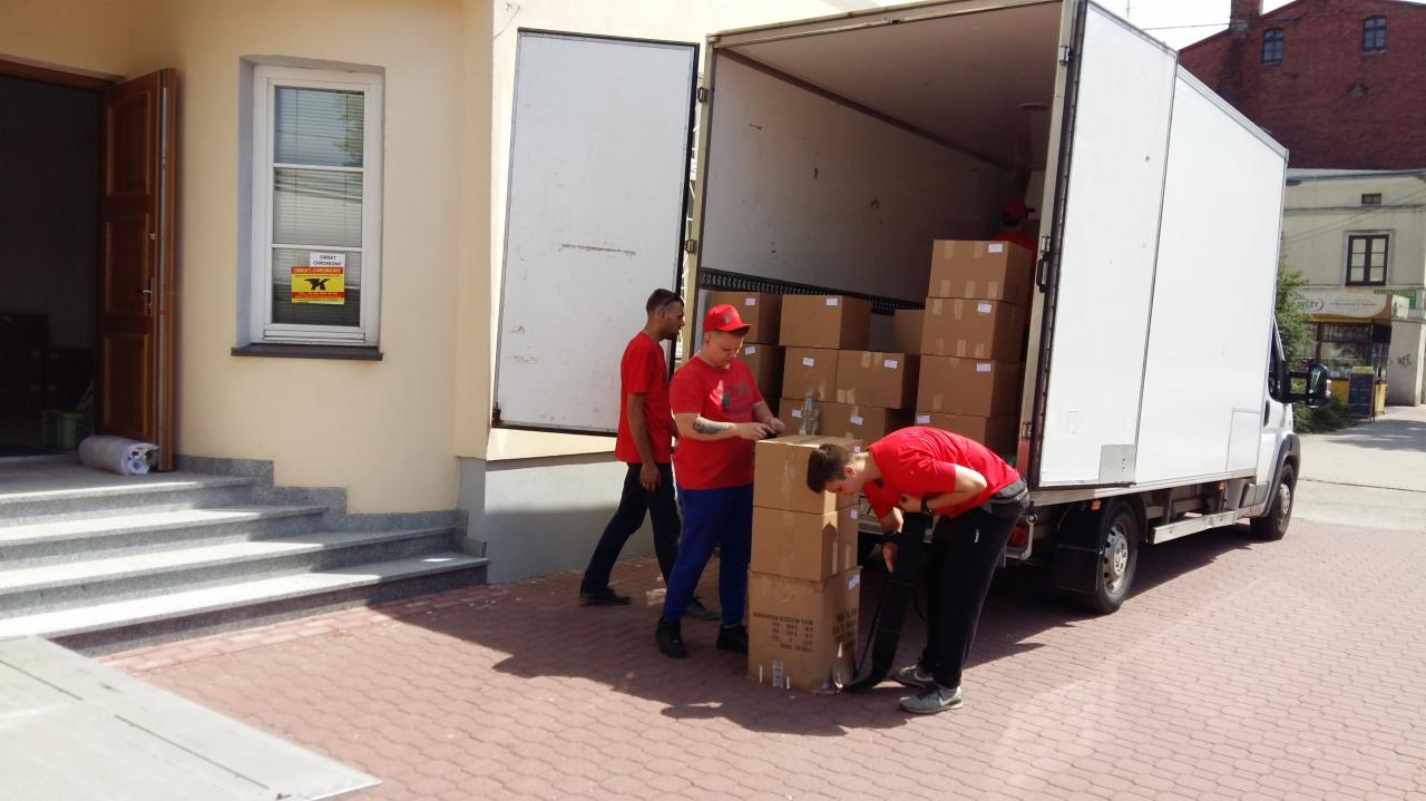 Trzech mężczyzn w czerwonych koszulkach wyładowuje kartony z białej furgonetki zaparkowanej przed budynkiem z szarymi schodami i oknem z żółtą naklejką.