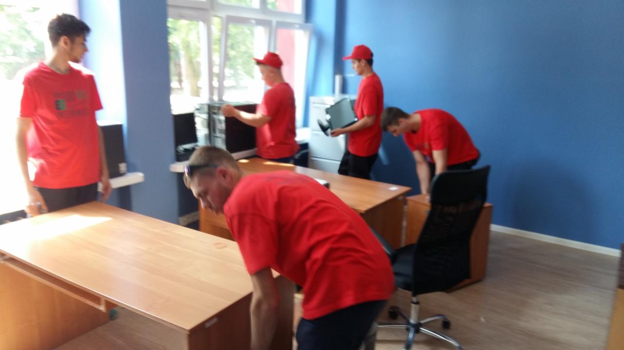 Zespół pracowników w czerwonych koszulkach przenosi biurka, komputery i drukarki w przestronnym biurze z niebieskimi ścianami podczas relokacji firmy.