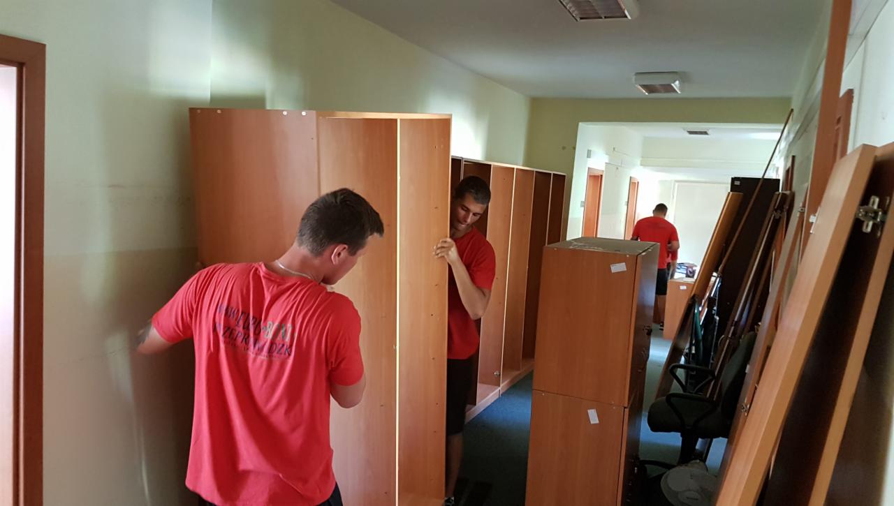 Dwóch mężczyzn w czerwonych koszulkach przenosi drewniane regały w korytarzu biurowym podczas relokacji.