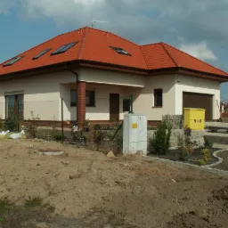 Parterowy dom jednorodzinny z czerwonym dachem, widoczne okna dachowe i garaż. Teren wokół domu częściowo zagospodarowany, z widocznymi elementami ogrodu.