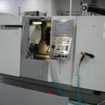 Biała tokarka CNC z otwartą komorą roboczą, widoczny panel sterowania i uchwyt narzędziowy. Oświetlenie górne, neutralne tło warsztatowe.