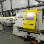 Biało-żółta maszyna CNC z widocznym ekranem sterowania w hali produkcyjnej, z elementami instalacji pod sufitem i czerwoną skrzynką narzędziową w tle.