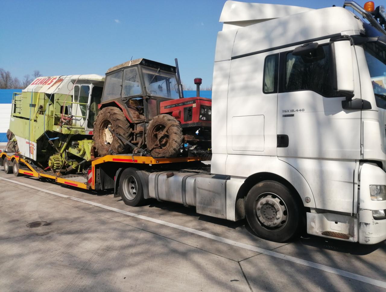 Ciężarówka MAN TGX 18.440 przewozi na niskopodwoziowej platformie kombajn zbożowy i traktor Zetor Turbo, widok z boku na betonowym placu.