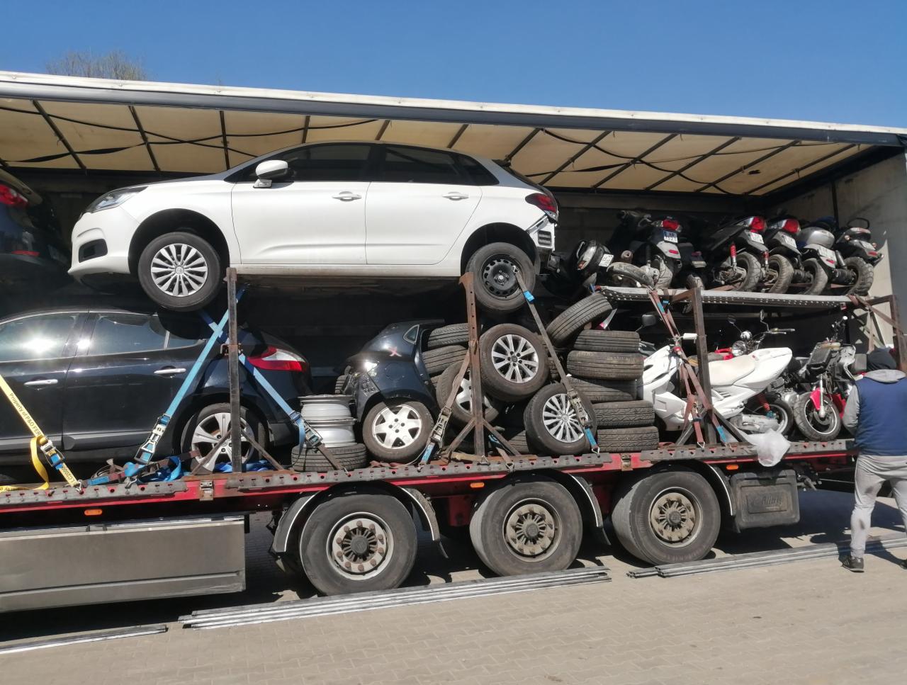 Wielopoziomowa laweta załadowana samochodami osobowymi, motocyklami i oponami, zabezpieczona pasami transportowymi, widok z boku.