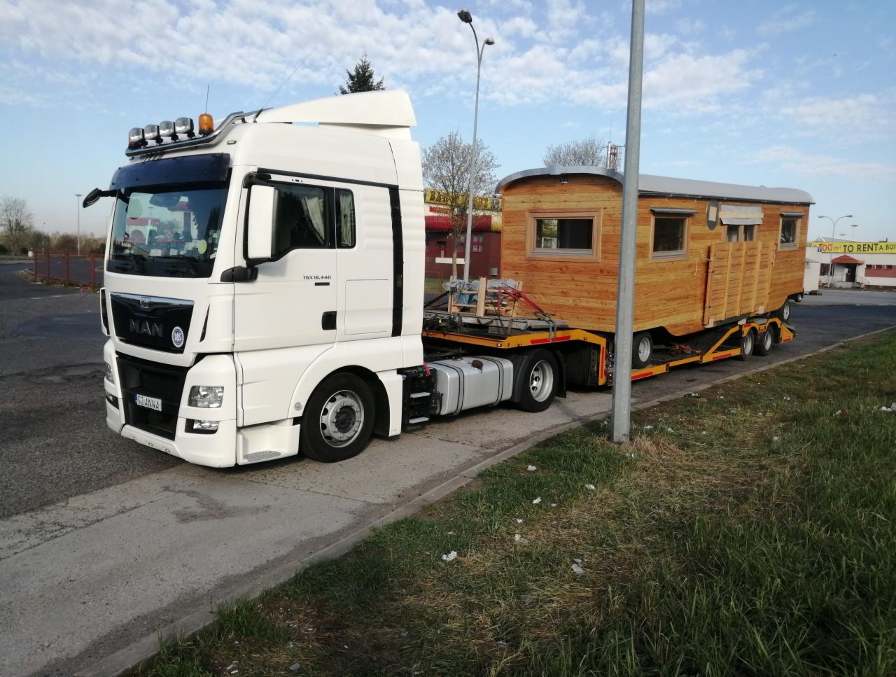 Biała ciężarówka MAN z naczepą niskopodwoziową przewożąca drewniany, mobilny domek z oknami, zaparkowana na parkingu.