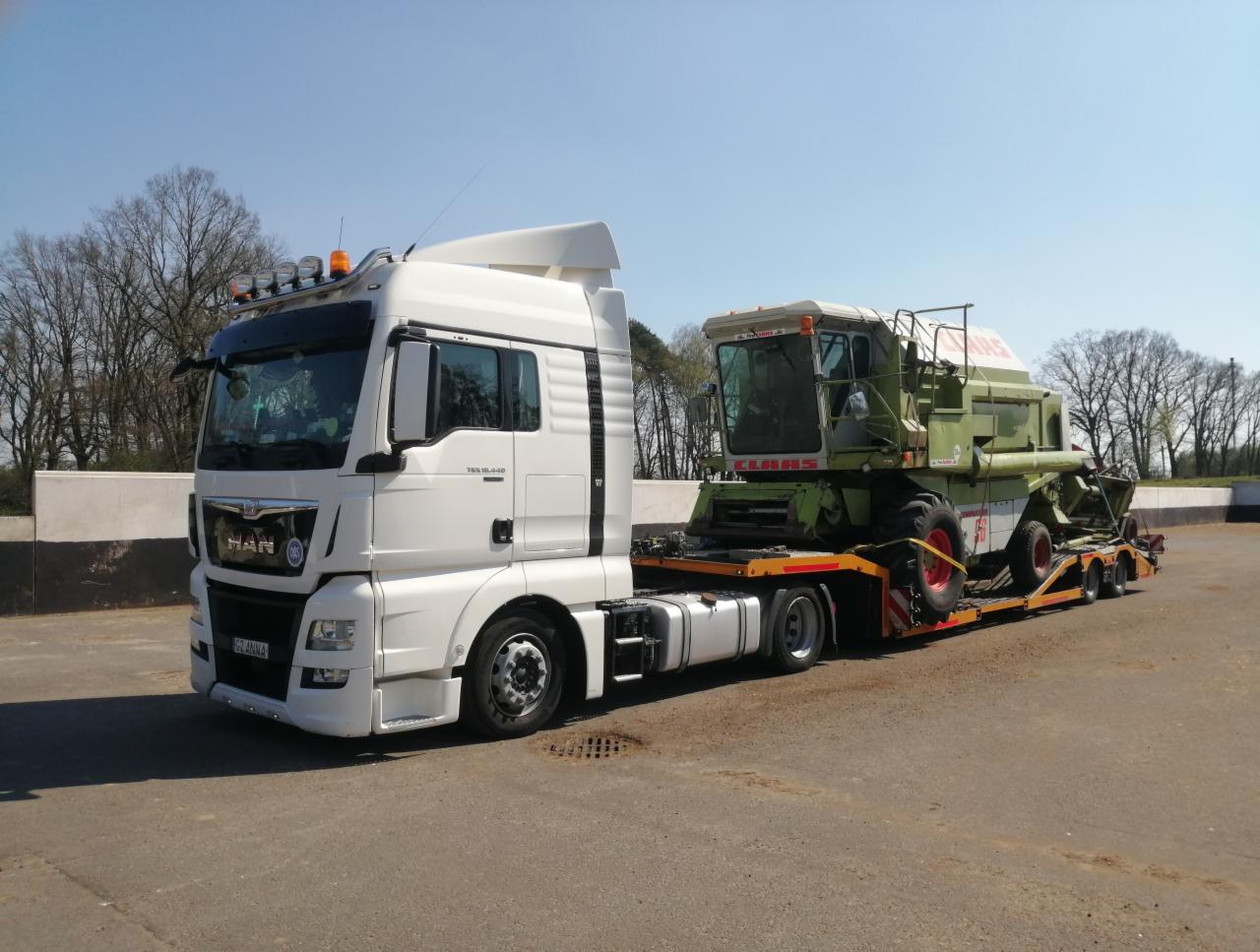 Biała ciężarówka MAN z naczepą niskopodwoziową przewożąca zielony kombajn zbożowy marki Claas na betonowym placu w słoneczny dzień.