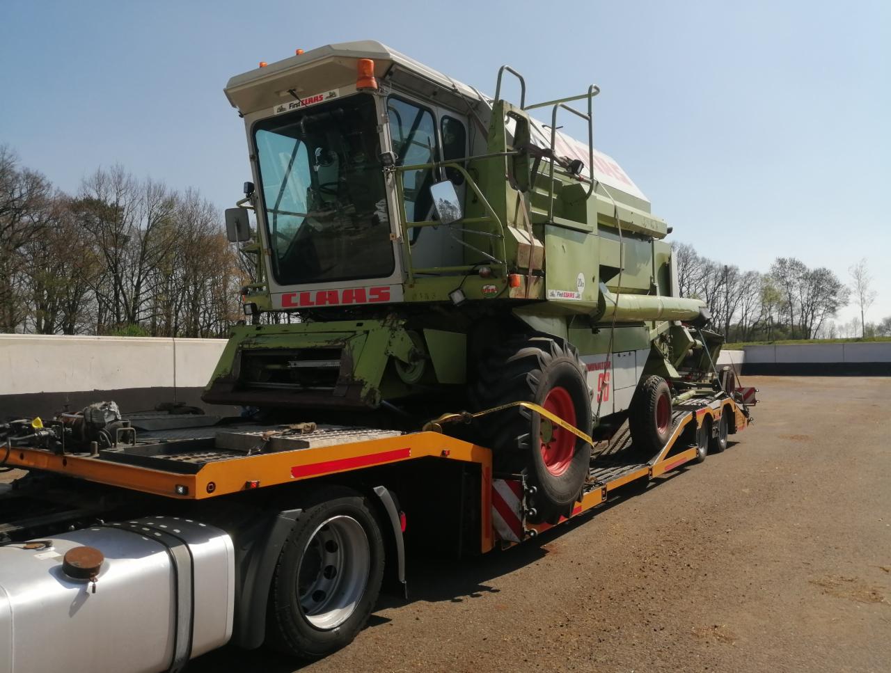 Zielony kombajn zbożowy marki Claas, zabezpieczony żółtymi pasami, przewożony na niskopodwoziowej lawecie. Widoczne opony z czerwonymi felgami i elementy mocujące. W tle drzewa i błękitne niebo.