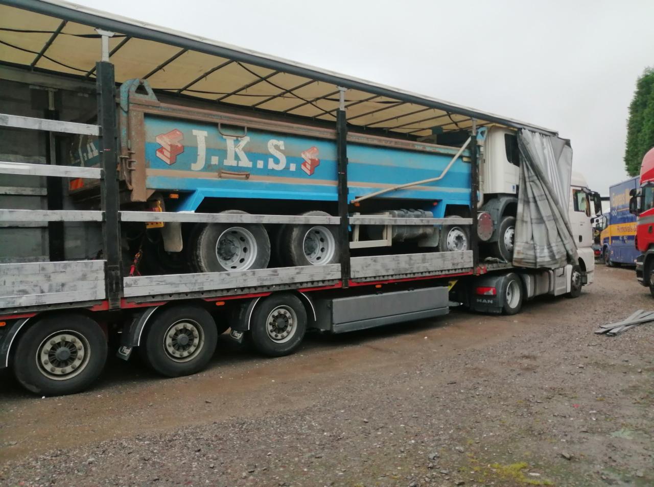 Ciężarówka MAN przewozi na platformie niebieską skrzynię ładunkową z napisem J.K.S. na tle szarego nieba.