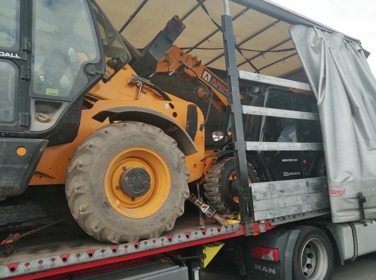 Pomarańczowa ładowarka teleskopowa zabezpieczona pasami transportowymi na platformie ciężarówki z plandeką.