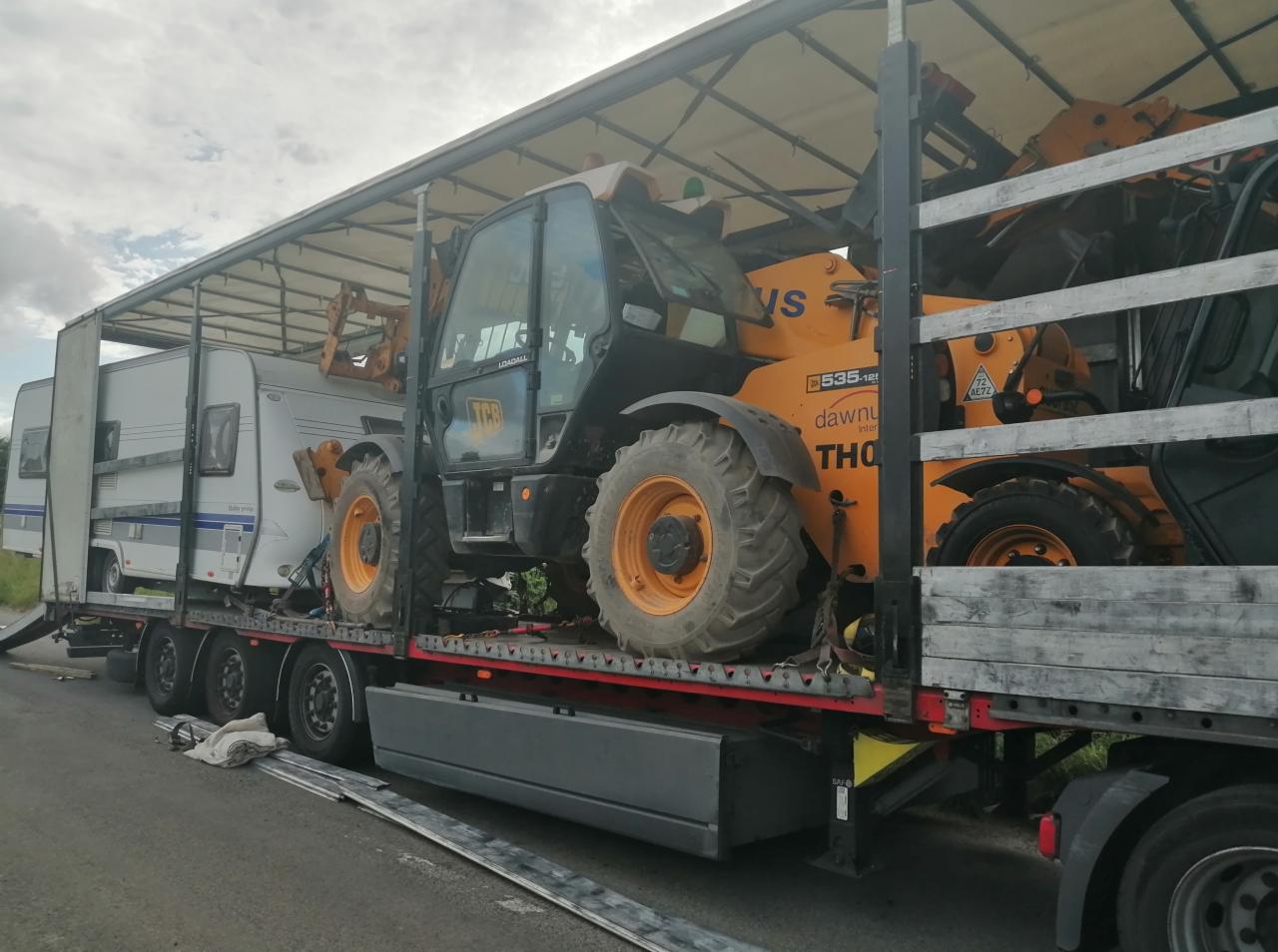 Nisko zawieszona laweta z plandeką przewozi jednocześnie przyczepę kempingową i dwie pomarańczowe ładowarki teleskopowe.