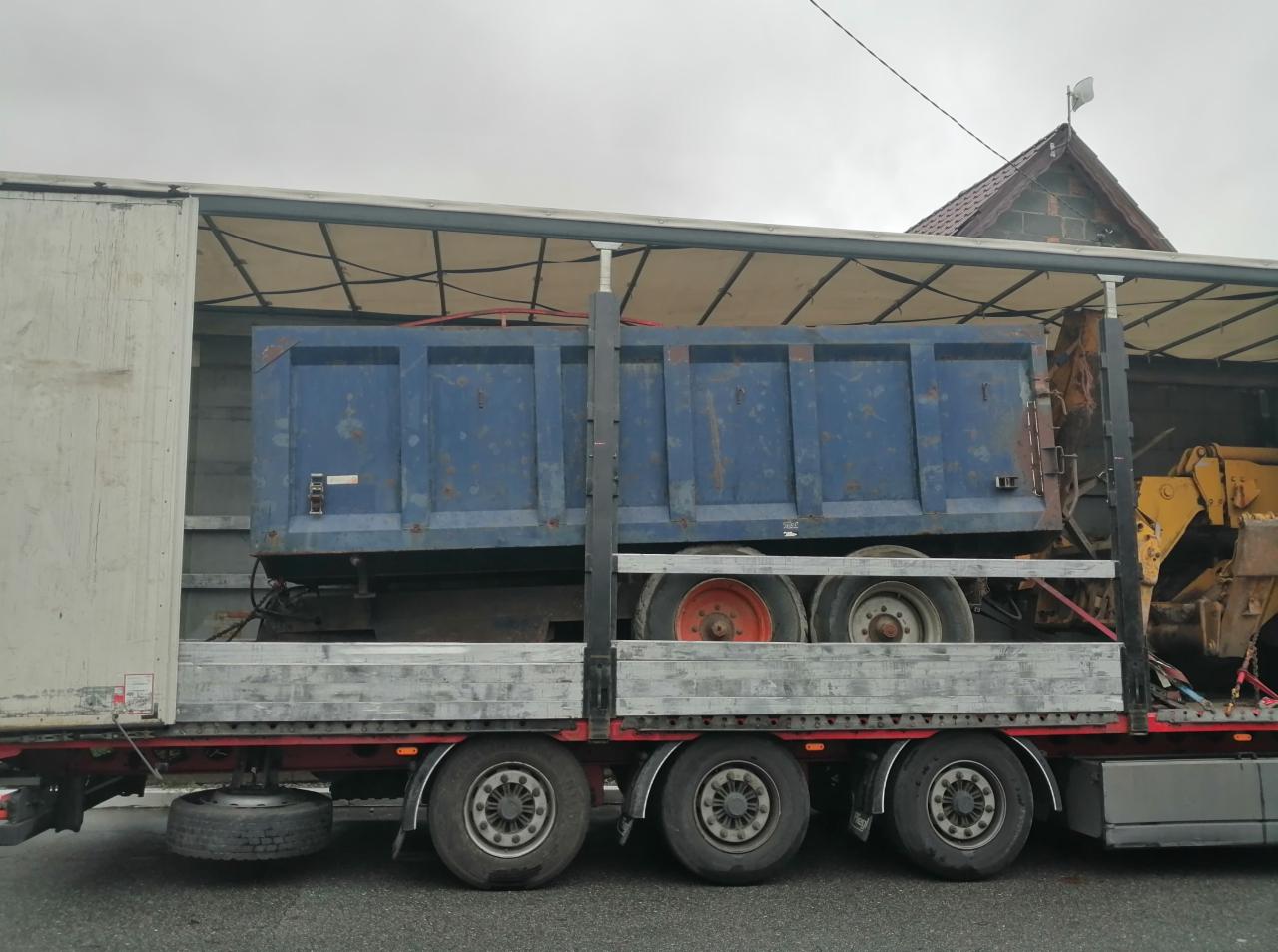 Ciężarówka z naczepą przewożąca niebieską skrzynię ładunkową i żółtą koparkę, widoczne koła i podwozie, szary dzień.