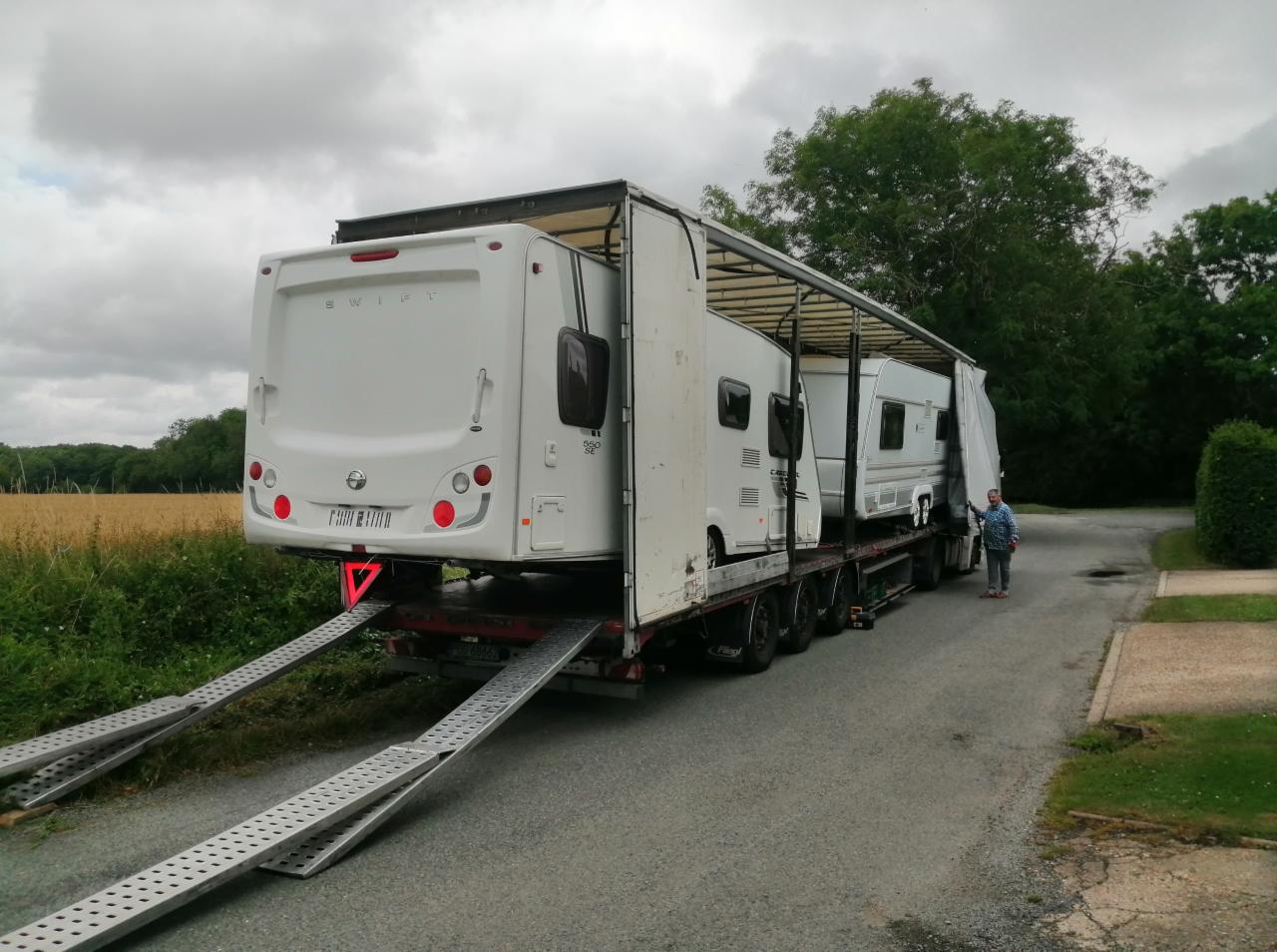 Dwa białe kampery marki Swift załadowane na niskopodwoziową lawetę z rampami, na tle pola i drzew