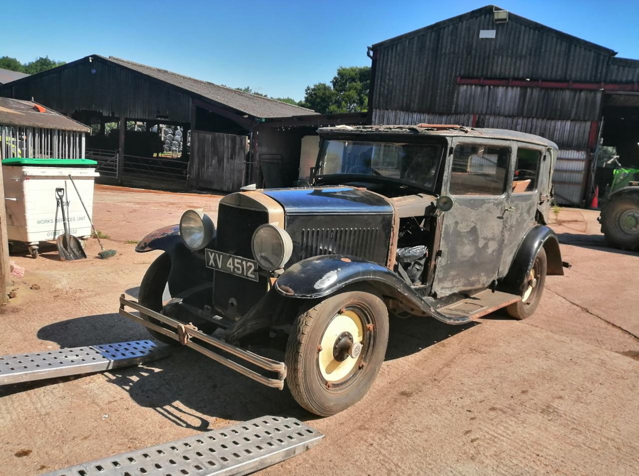 Zardzewiały czarny samochód osobowy z numerem rejestracyjnym XV 4512, częściowo rozebrany, na rampie w wiejskim otoczeniu z budynkami gospodarczymi w tle.