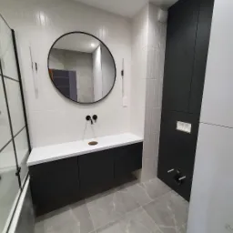 Nowoczesna łazienka z czarnymi elementami: szafka pod umywalkę, okrągłe lustro i kabina prysznicowa z czarnymi ramami. Białe płytki na ścianie i szare płytki na podłodze.