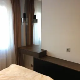 Nowoczesna szafka z lustrem i wiszącą lampą, ciemne drewno, czarne szuflady, beżowa pościel, brązowe zasłony.