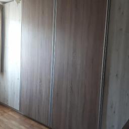 AS line - Zamknięta szafa wnękowa z przesuwnymi drzwiami o drewnopodobnym wzorze, z widocznymi szynami i fragmentem okna z zasłoną w tle.