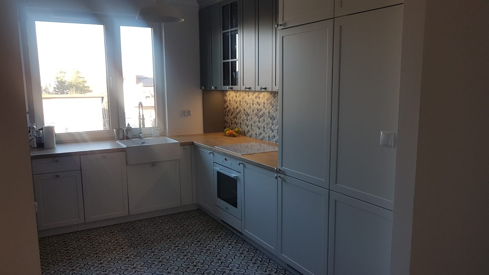 Jasna kuchnia z białymi frontami, drewnianym blatem i wzorzystą podłogą, widok na okno z zlewozmywakiem.