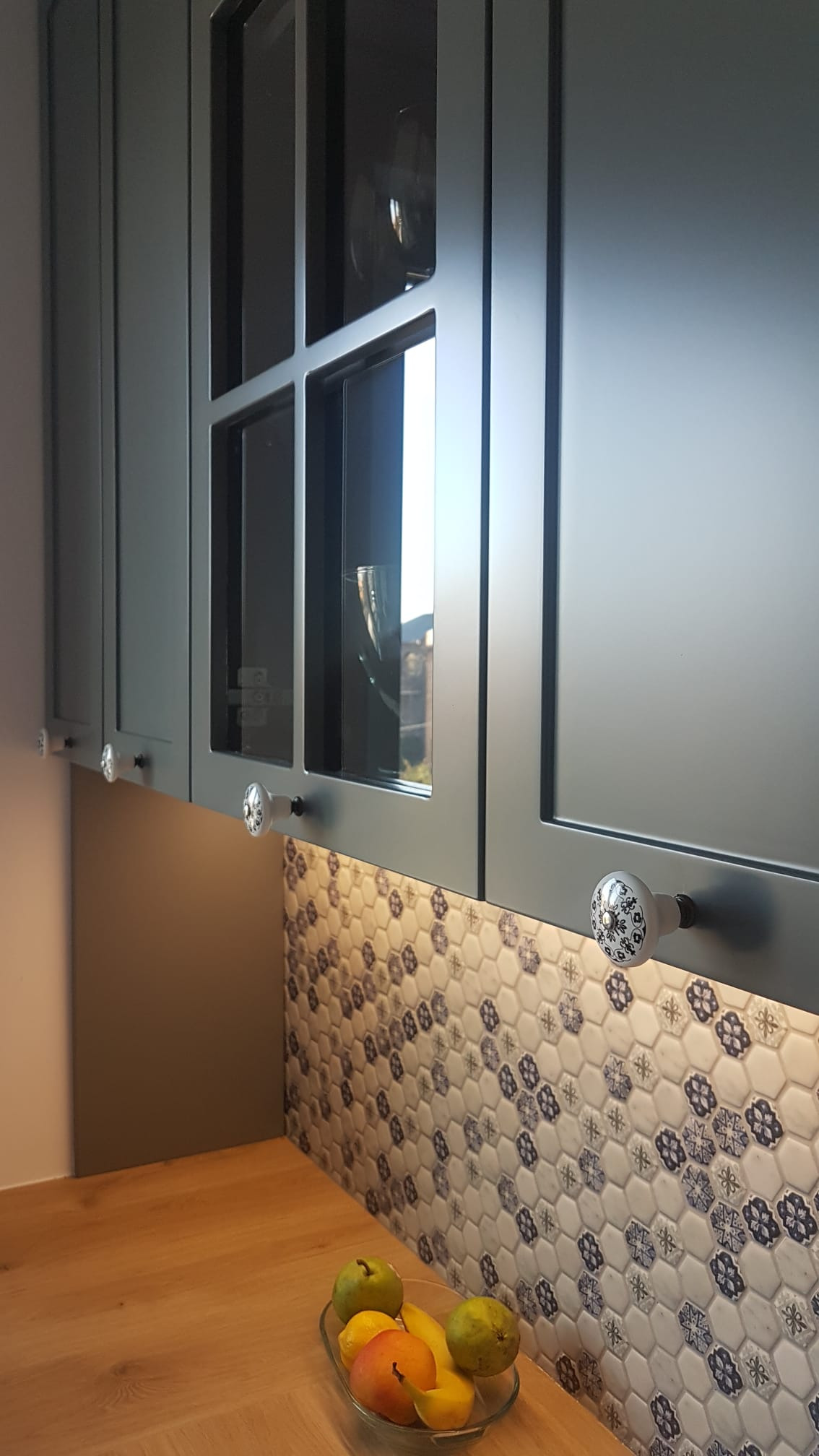 Szare szafki kuchenne z przeszklonymi frontami, ozdobnymi gałkami i heksagonalnymi płytkami na ścianie, oświetlone listwą LED, z owocami w szklanej misie na blacie.