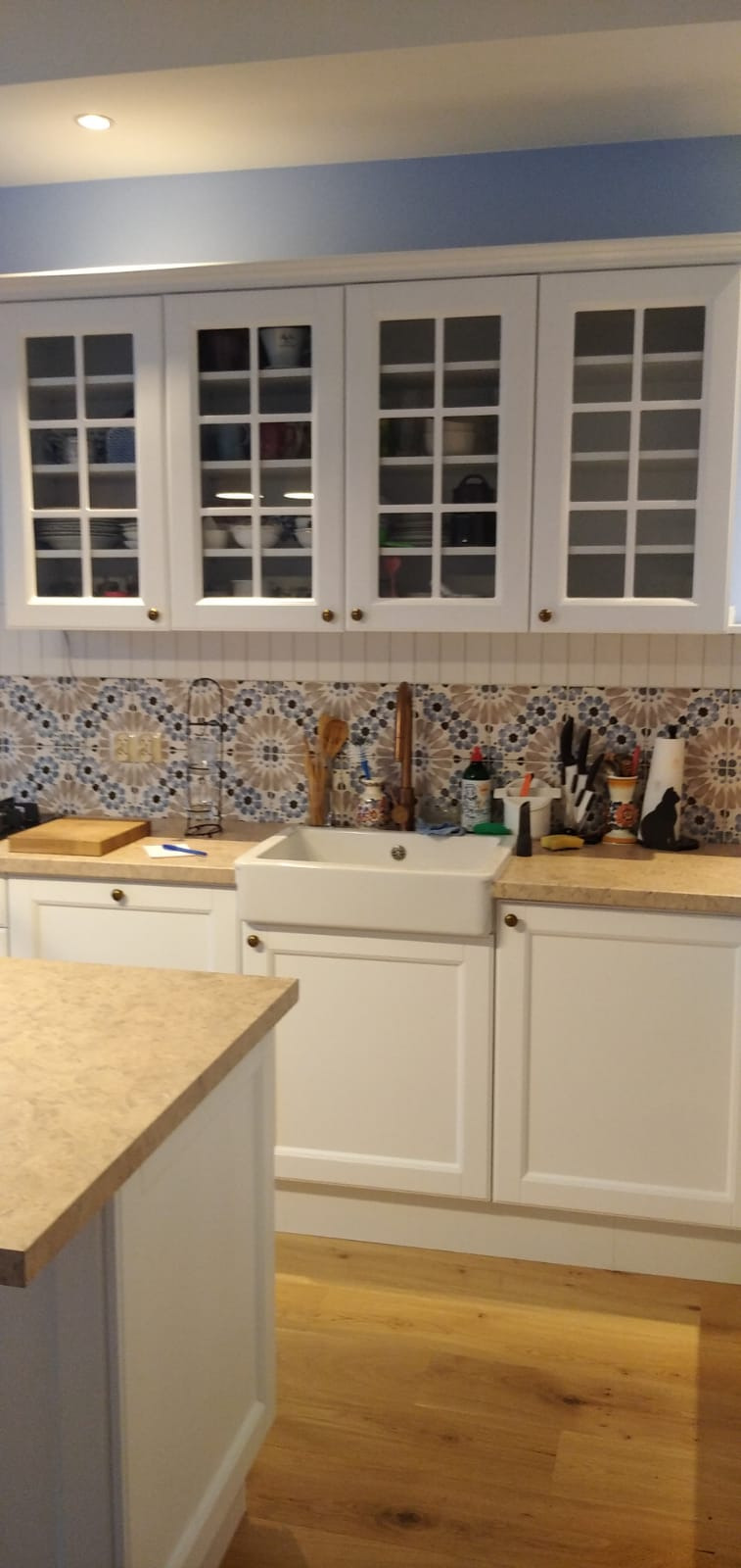 Białe szafki kuchenne z przeszklonymi frontami, ceramiczny zlew typu farmhouse i mozaikowa ściana nad blatem w kuchni z wyspą.