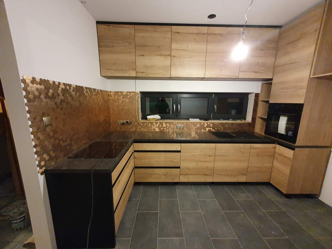 Jasna kuchnia z drewnopodobnymi frontami szafek, czarnym blatem i miedzianą mozaiką heksagonalną na ścianie, widoczny fragment podłogi z szarych płytek.