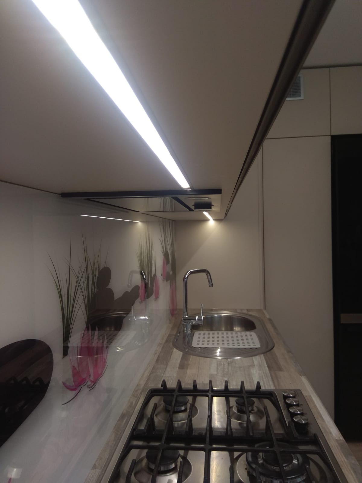 Nowoczesna kuchnia z blatem imitującym drewno, szklanym panelem z motywem roślinnym i oświetleniem LED pod szafkami.